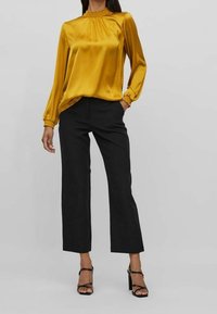 Blouse en satin moutarde avec col et poignets froncés, associée à un pantalon large noir et des sandales à talons hauts noires à lanières.