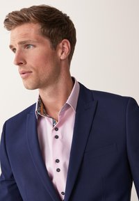 Chaqueta de traje azul marino con una camisa de textura rosa que presenta un interior del cuello con estampado y botones oscuros. Primer plano del escote y el detalle del hombro.
