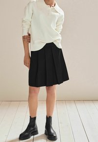 Cremefarbener Strickpullover mit Kragen und Knopfverschluss, kombiniert mit einem schwarzen plissierten Rock und schwarzen Stiefeletten auf einem Holzboden.