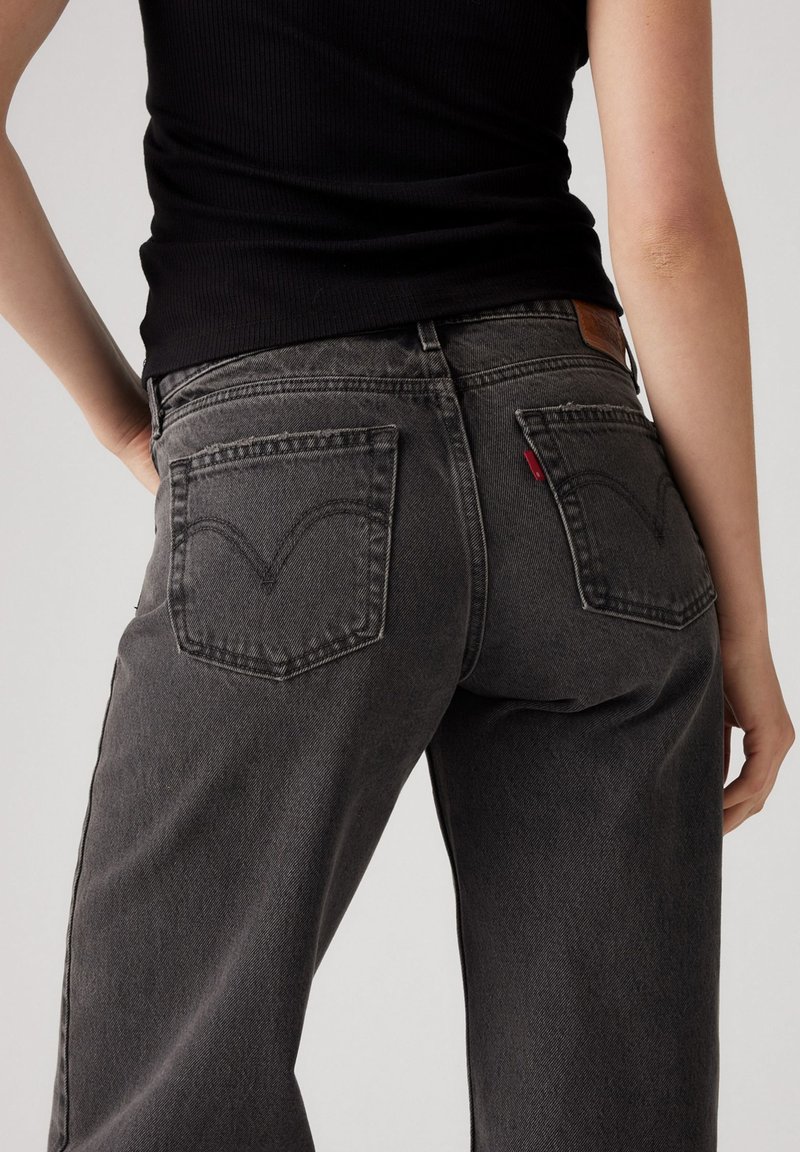 Jeans de mezclilla gris con un diseño de cintura alta, que cuentan con dos bolsillos traseros, un parche de cuero y un detalle de etiqueta roja en el bolsillo derecho.