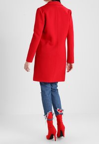 Rød uldfrakke med lige snit, lange ærmer og krave, kombineret med slidte blå jeans og røde stiletstøvler.