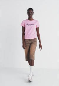 Camiseta de algodón rosa con un diseño gráfico, combinada con shorts de mezclilla marrón claro. El modelo lleva calcetas blancas y zapatillas grises. Fondo neutro.