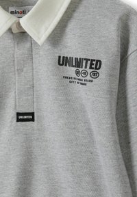 Szary polo wykonany z bawełny z białym kołnierzykiem. Zawiera czarny napis "UNLIMITED" oraz szczegóły logo. Posiada czarną metkę na dolnym końcu.