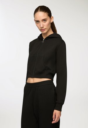 Kobieta z związanymi włosami, ubrana w czarną, krótką bluzę z zamkiem i pasujące czarne dresy, stojąca na tle jasnego, jednolitego tła.