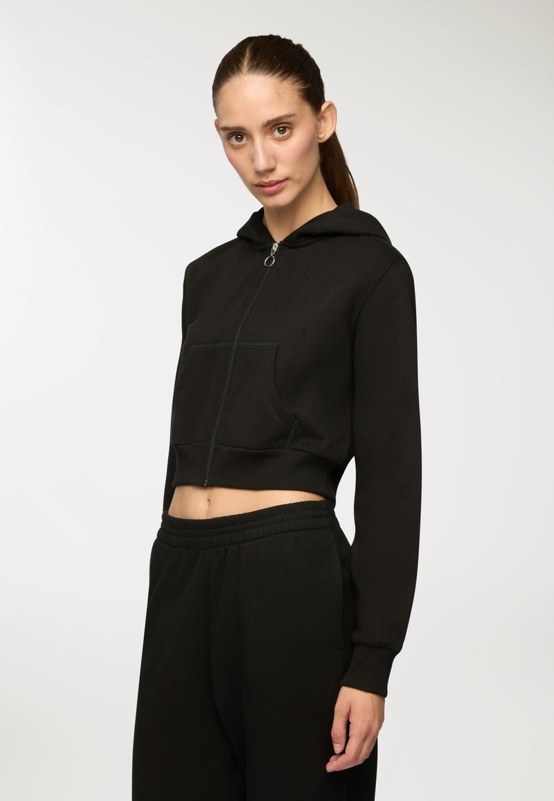 Donna con i capelli raccolti, che indossa un hoodie nero a zip corto e pantaloni della tuta neri abbinati, in piedi su uno sfondo chiaro e semplice.