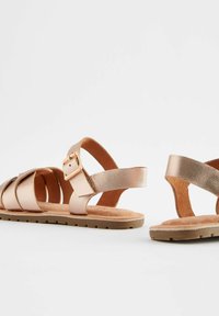 Sandali in oro rosa metallico con una finitura testurizzata, caratterizzati da molteplici cinturini intrecciati e una fibbia. Suola morbida e design piatto.