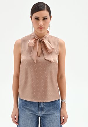 Donna che indossa una camicetta beige senza maniche con piccoli pois bianchi e colletto con fiocco, abbinata a jeans blu e braccialetto d'argento.