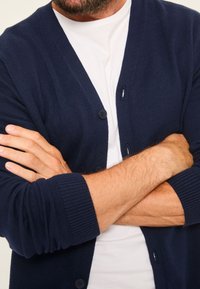 Mann verschränkt die Arme, trägt eine dunkelblaue, geknöpfte Strickjacke über einem weißen T-Shirt, zeigt Unterarme und einen Teil des Bartes.