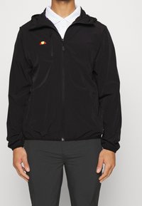 Chaqueta ligera negra con cierre de cremallera en el frente, puños elásticos y capucha ajustable. Presenta un pequeño logo rojo y amarillo en el pecho.