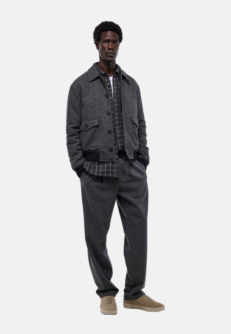Veste en laine grise avec fermeture à boutons et poches avant, portée par-dessus une chemise à carreaux noir et blanc, accompagnée de pantalons gris ample.