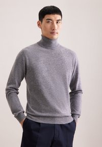 Grauer Rollkragenpullover mit langen Ärmeln, aus weichem Strickmaterial; mit gerippten Bündchen und Saum, getragen mit navyfarbenen Hosen.