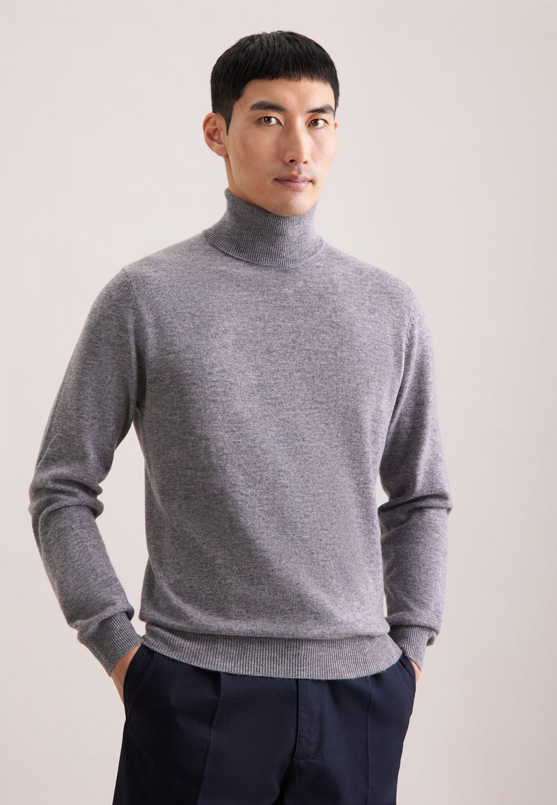 Grauer Rollkragenpullover mit langen Ärmeln, aus weichem Strickmaterial; mit gerippten Bündchen und Saum, getragen mit navyfarbenen Hosen.