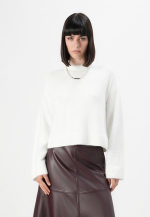 Vrouw met stijl donker haar draagt een witte pluizige trui, parelketting en een hooggeplaatste donkerbruine leren rok, staande tegen een witte achtergrond.