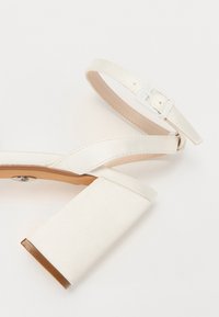 Λευκά σανδάλια με τετράγωνο τακούνι και γυαλιστερή φινίρισμα, που διαθέτουν φαρδύ λουρί πάνω από τα δάχτυλα και ρυθμιζόμενο λουρί στον αστράγαλο με μεταλλική αγκράφα.