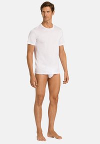 Bílá bavlněná tričko s krátkými rukávy, kulatým výstřihem a přiléhavým střihem. Model nosí bílé trenýrky, stojí bosý na jednobarevném pozadí.