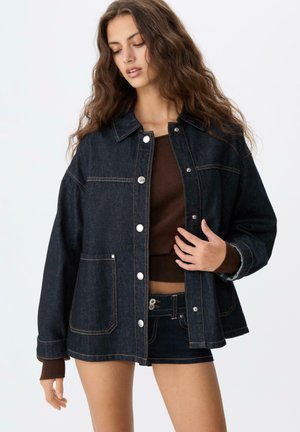 Veste en denim foncé avec col, boutons-pression et poches. Présente des coutures contrastées et une coupe décontractée. Top marron intérieur visible.