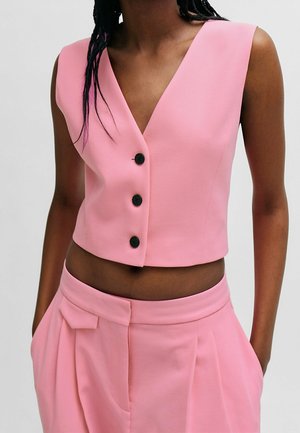 Kobieta ubrana w różowy bezrękawnik z czarnymi guzikami i dopasowane różowe spodnie z wysokim stanem, ręce w kieszeniach, na neutralnym tle.