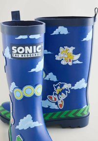 Modré holínky Sonic the Hedgehog s obrázky Sonica, Tailse, mraků, prstenců a zelenými chevronovými pruhy blízko podrážek.