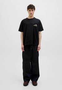 Černé bavlněné tričko s krátkým rukávem, na kterém se nachází malé, barevné logo "ATELIER ORIGINAL" na hrudi, v kombinaci s volnými, tmavými džíny.