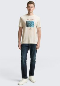 Beżowa bawełniana koszulka z niebieskim nadrukiem, krótki rękaw, okrągły dekolt, w zestawie z ciemnoniebieskimi dżinsami i białymi trampkami.