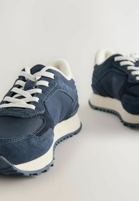 Tumedate sneakers, mis on valmistatud sametist ja siledast materjalist, valgete paeltega ja tekstureeritud kummist tallaga. Ümar varvas ja pehmendatud pahkluutoe osakaal.