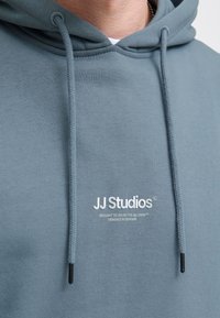 Fleece hoodie i dæmpet teal med snoretræk, med et hvidt trykt logo på brystet og en glat tekstur.