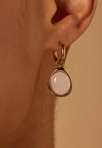 Gouden oorbel met een druppelvormige, ondoorzichtige roze steen. Gladde oppervlakte met een glanzende afwerking, hangt aan een eenvoudige sluiting.