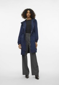 Marineblauwe waterdichte parka met een capuchon, elastische tailleband, rits- en knoopsluiting en twee voorkruiden. Gecombineerd met grijze wijde broek.