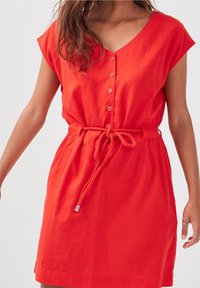 Femme portant une robe rouge à manches courtes avec des boutons sur le devant et une ceinture en corde nouée à la taille sur un fond uni.