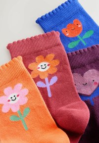 Quatre paires de chaussettes tricotées colorées : orange avec une fleur rose, rose avec une fleur souriante, bordeaux avec un visage en forme de cœur, bleu avec une tulipe souriante.