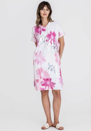 Vrouw draagt een witte hoodie-jurk tot op de knie met roze bloemmotieven en zilveren tekst, gecombineerd met witte slidesandalen, staand tegen een effen achtergrond.