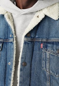 Uomo che indossa una giacca di jeans blu con fodera in sherpa bianca sopra un hoodie grigio chiaro, con il tag rosso Levi's visibile sulla tasca del petto.