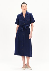 Vestido de lino azul marino con mangas cortas, botones en el frente, dos bolsillos en el pecho y detalle de cintura ajustable. Escote cuadrado y bajo recto.