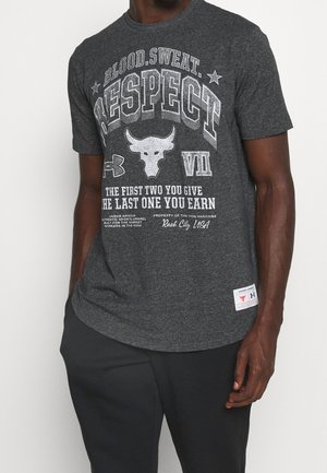 Mann trägt ein dunkelgraues T-Shirt mit dem Text "Blood. Sweat. Respect" und Bullenlogo, kombiniert mit schwarzen Hosen.
