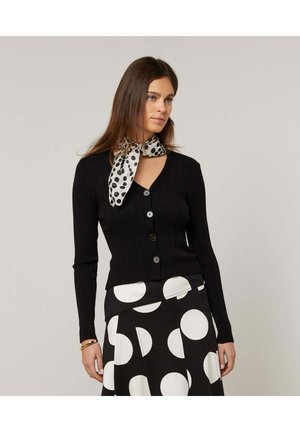 Vrouw draagt een zwarte geribbelde cardigan met knopen, een zwarte rok met grote witte polka dots en een witte sjaal met polka dots om haar nek, staand tegen een grijze achtergrond.