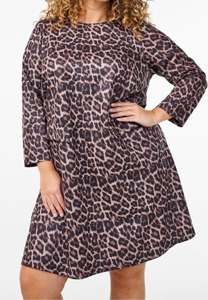 Kvinde iført en langærmet, knælængde kjole med leopardprint med hånden på hoften mod en hvid baggrund.