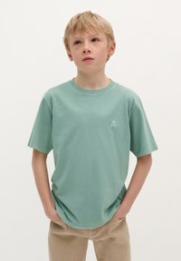 T-shirt en coton vert clair avec des manches courtes, col rond et un petit graphique de crâne sur la poitrine gauche. Texture lisse, coupe décontractée.
