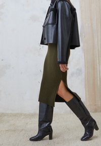 Svart kort skinnjacka över en ribbad olivgrön midiklänning med sidoklipp, tillsammans med knähöga svarta klackstövlar.