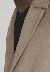 Primer plano de una persona que lleva un abrigo de lana beige sobre un suéter negro, mostrando la solapa del abrigo y parte del escote.