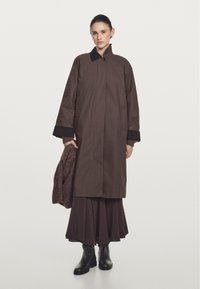Brown raincoat with a high collar, front button closure, and side pockets. Worn over a long brown skirt and holding a knitted sweater. Black ankle boots.