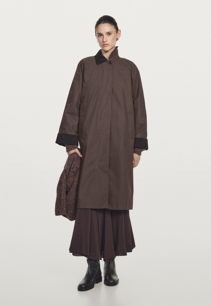 Brown raincoat with a high collar, front button closure, and side pockets. Worn over a long brown skirt and holding a knitted sweater. Black ankle boots.