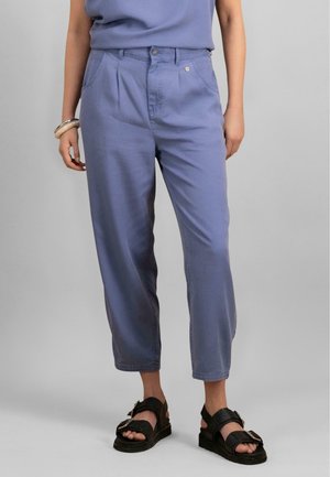Pantalons violet clair en coupe décontractée, avec une taille haute, deux poches avant et une texture lisse. Portés avec des sandales noires.