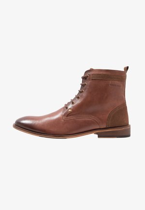 Botte en cuir brun à lacets, avec un bout rond et un panneau arrière texturé. Elle présente des œillets dorés et une semelle en caoutchouc.