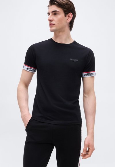 Jeune homme portant un t-shirt noir Moschino avec des bandes logo aux bords des manches et un pantalon noir assorti avec des bandes blanches sur les côtés.