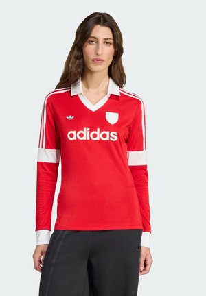 Jersey rojo de manga larga con cuello y mangas blancas, que presenta tres rayas blancas en los hombros y un gran logo de "adidas" en el centro.