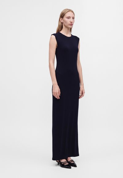 Femme debout, face à l’objectif, portant une robe longue noire sans manches et des chaussures noires à bout pointu sur fond blanc uni.