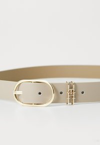 Beige leren riem met een gouden ovale gesp. Inclusief gouden hardware accenten en gaatjes voor aanpassing. Gladde textuur, minimalistisch ontwerp.