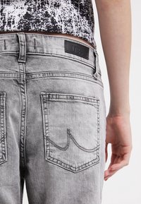 Jeans de mezclilla gris con una textura lavada única, bolsillos traseros dobles con acentos de costura y una etiqueta en la cinturilla.