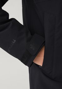 Close-up van een hand in een zwarte jaszak, waarbij de mouw met verstelbare klittenbandsluiting en subtiele logo-details zichtbaar is.