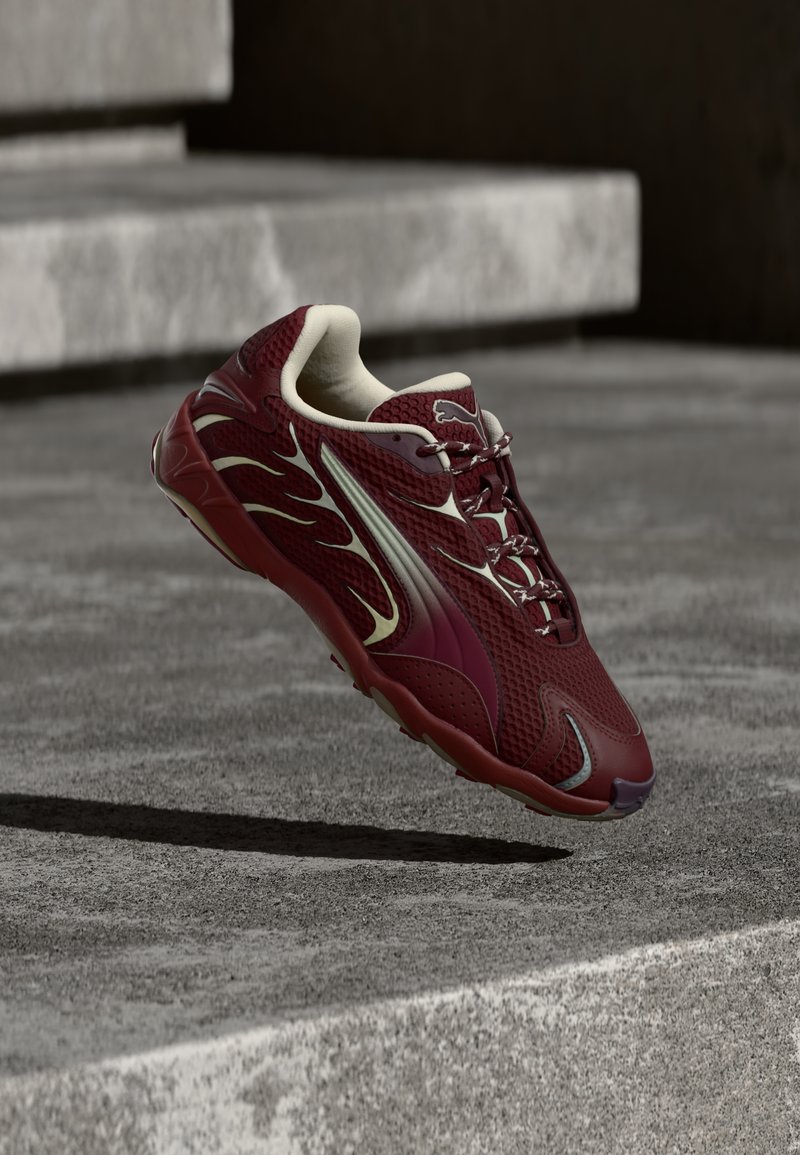 Burgundi Puma sneaker fehér részletekkel, ami a betonlépcsők felett lebeg, árnyékot vetve a lent lévő földre.